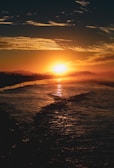 A vibrant sunset casting a golden glow over the ocean, with silhouettes of distant trees and mountains on the horizon. The sky is adorned with textured clouds reflecting the warm hues of the setting sun.