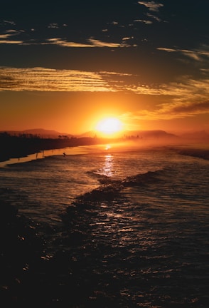 A vibrant sunset casting a golden glow over the ocean, with silhouettes of distant trees and mountains on the horizon. The sky is adorned with textured clouds reflecting the warm hues of the setting sun.
