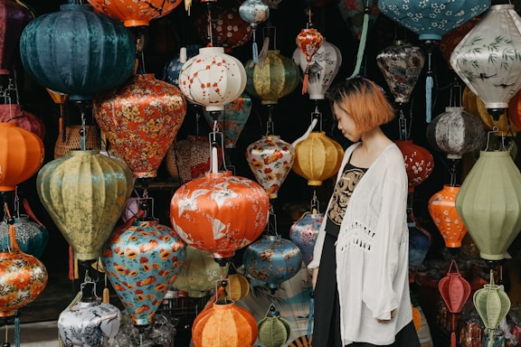 A woman with short hair and a white jacket stands next to a display of colorful paper lanterns. The lanterns are of various shapes, sizes, and patterns, hanging closely together.