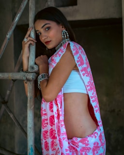 A young woman wearing a white crop top and a pink and white patterned sari. She is standing beside a bamboo structure, adorned with large earrings and bracelets. Her expression is poised and contemplative.