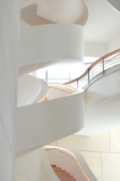 A modern, spiral staircase with sweeping white curves and wooden steps, illuminated by soft natural light coming through large windows on the upper level. The design is minimalist, with clean lines and smooth, flowing shapes.