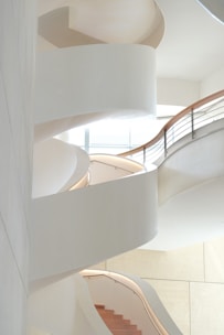 A modern, spiral staircase with sweeping white curves and wooden steps, illuminated by soft natural light coming through large windows on the upper level. The design is minimalist, with clean lines and smooth, flowing shapes.