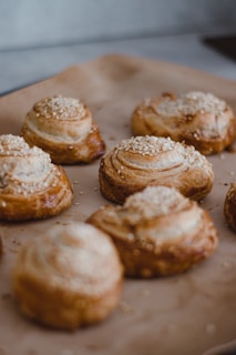 A close-up view of several freshly baked pastries topped with sesame seeds, arranged on a baking tray lined with parchment paper. The pastries have a golden-brown crust with visible layers of flaky dough.