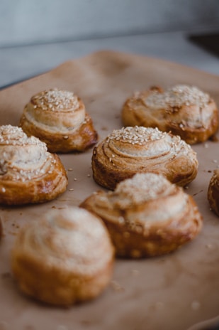 A close-up view of several freshly baked pastries topped with sesame seeds, arranged on a baking tray lined with parchment paper. The pastries have a golden-brown crust with visible layers of flaky dough.