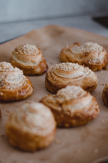 A close-up view of several freshly baked pastries topped with sesame seeds, arranged on a baking tray lined with parchment paper. The pastries have a golden-brown crust with visible layers of flaky dough.
