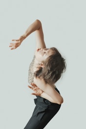 A person is captured mid-dance in an expressive pose, with their arm raised and head tilted back. They are wearing a patterned sleeveless top and black pants, and their hair appears to be tousled. The background is plain and light-colored.