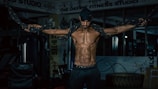 A muscular man is exercising in a gym, holding heavy ropes with his arms extended. The environment features fitness equipment and motivational posters, creating a focused and intense atmosphere.