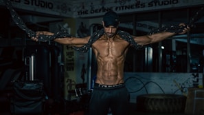 A muscular man is exercising in a gym, holding heavy ropes with his arms extended. The environment features fitness equipment and motivational posters, creating a focused and intense atmosphere.