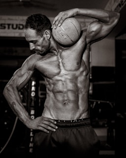 A muscular man is holding a basketball against his shoulder, showcasing a defined physique. The scene is captured in a gym setting with dim lighting, creating strong shadows and highlights on the man's body. The image is in black and white, adding a dramatic and intense effect.
