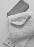 A neatly folded, soft-textured sweater rests on top of a magazine with the word 'REAL' visible. Positioned beside it is a sleek, gray leather pouch embossed with a brand name.