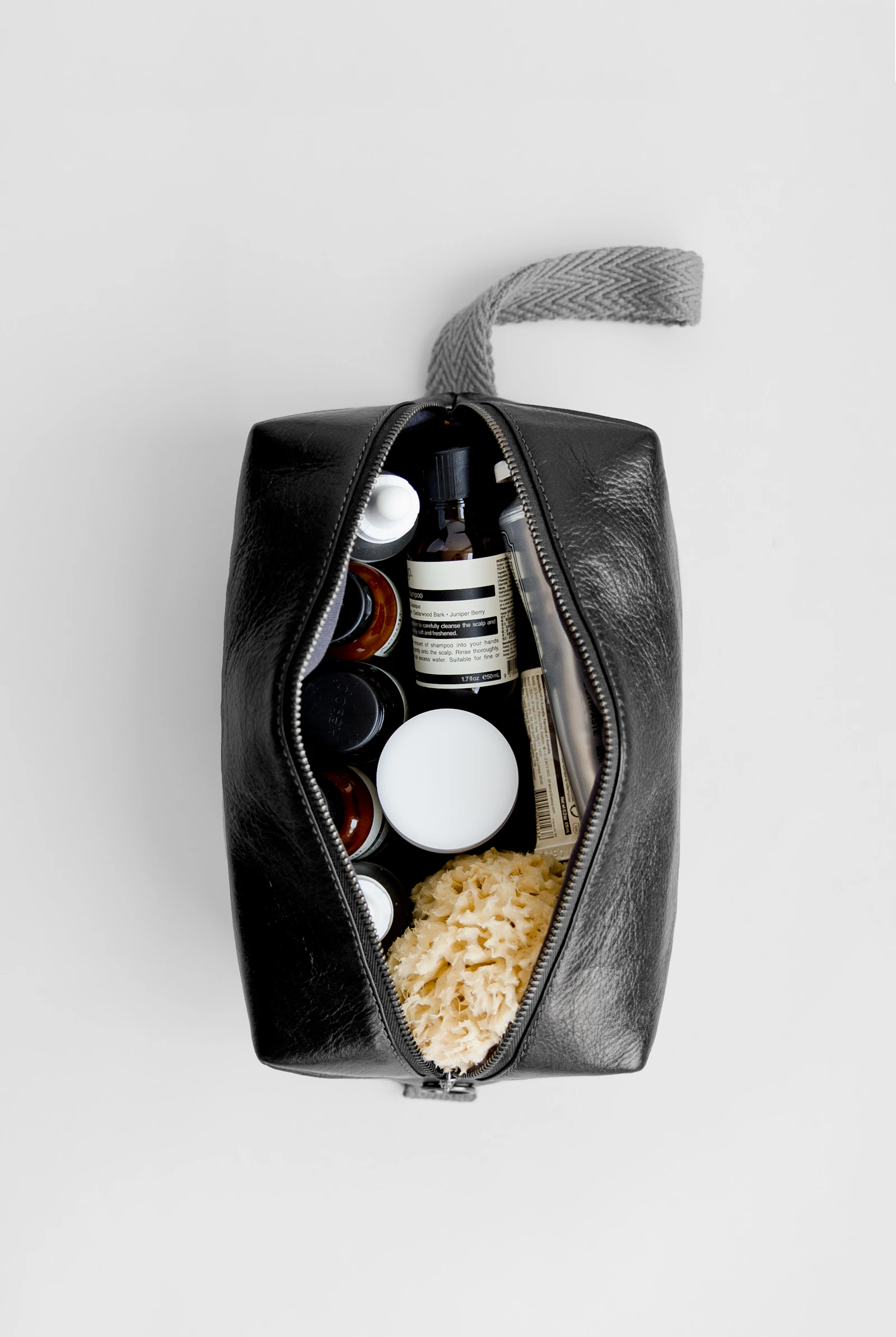 Photograph showing open black toiletry pouch holds small bottles, tubes, a jar and a natural sponge.