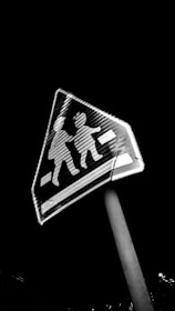 A triangular pedestrian crossing sign with silhouettes of two children, displayed at an angle. The sign has a high contrast appearance against a dark night sky.