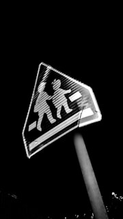 A triangular pedestrian crossing sign with silhouettes of two children, displayed at an angle. The sign has a high contrast appearance against a dark night sky.