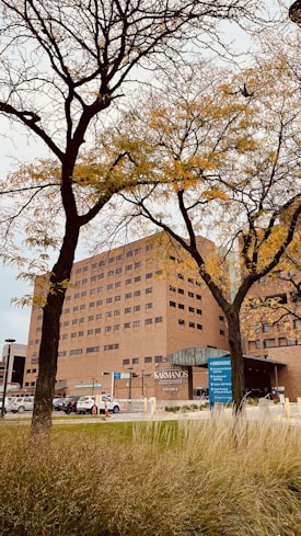 A large brick building with multiple windows stands behind tall trees with sparse branches, likely in the autumn season. The building features a prominent sign indicating 'Karmanos Cancer Institute' and there are parking and emergency signs visible near the entrance.