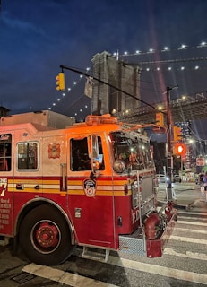 A fire truck is parked on a city street at night, with its lights on. The Brooklyn Bridge is illuminated in the background under a deep blue evening sky. Traffic lights and buildings are visible, adding to the urban setting.