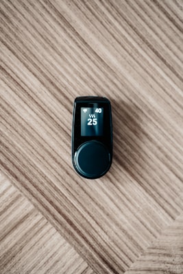 A sleek black electronic device with a digital display on top, showing numbers 40 and 25 along with icons, is placed on a wooden surface with a diagonal pattern.