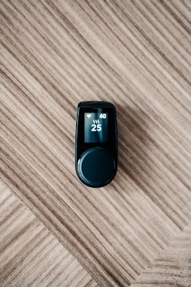 A sleek black electronic device with a digital display on top, showing numbers 40 and 25 along with icons, is placed on a wooden surface with a diagonal pattern.