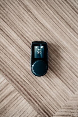 A sleek black electronic device with a digital display on top, showing numbers 40 and 25 along with icons, is placed on a wooden surface with a diagonal pattern.