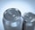 Two aluminum beverage cans are positioned closely together. The focus is on the top lids with pull tabs, showcasing a metallic and reflective surface. Lighting highlights the shiny texture and smooth curves of the cans.