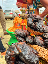 A bustling outdoor market scene with various goods displayed. In the foreground, there is a basket filled with smoked or roasted food items, possibly meat or fish. Behind them are colorful containers and baskets filled with dried foods or grains. A person is bending over, likely interacting with the goods, and there are other market stalls visible in the background along with parked cars.