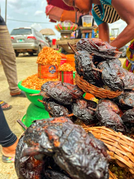 A bustling outdoor market scene with various goods displayed. In the foreground, there is a basket filled with smoked or roasted food items, possibly meat or fish. Behind them are colorful containers and baskets filled with dried foods or grains. A person is bending over, likely interacting with the goods, and there are other market stalls visible in the background along with parked cars.