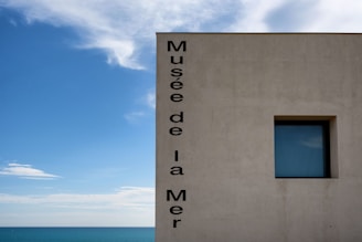 A minimalist building wall is adorned with vertical black text reading 'Musée de la Mer' next to a square window. The background features a bright blue sky with scattered white clouds and a view of the ocean meeting the horizon.