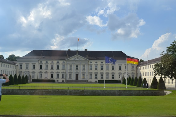 A large, classical-style white building with a symmetrical design, flanked by smaller wings on either side. The structure features numerous tall windows and a central pediment. Two flags, the European Union and German flags, are prominently displayed on flagpoles in front of the building. The foreground features a well-maintained green lawn bordered by a stone wall and hedges. A few people are visible, one taking a photo on the left.