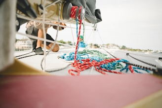 A person is kneeling on the deck of a sailboat, engaging in work or maintenance. Colorful ropes, mostly red and blue, are scattered across the boat's surface. The background shows a blurred view of what appears to be a marina or harbor.