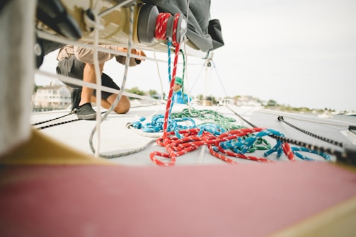 A person is kneeling on the deck of a sailboat, engaging in work or maintenance. Colorful ropes, mostly red and blue, are scattered across the boat's surface. The background shows a blurred view of what appears to be a marina or harbor.
