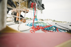 A person is kneeling on the deck of a sailboat, engaging in work or maintenance. Colorful ropes, mostly red and blue, are scattered across the boat's surface. The background shows a blurred view of what appears to be a marina or harbor.