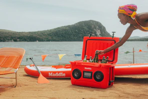 A portable wheeled cooler being pulled across a sandy beach with a family setting up a picnic nearby.