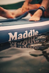 A close-up of a paddleboard with the brand name 'Maddle' visible. Feet are resting on the board, and a coiled leash is attached to an ankle. The water reflects the lettering on the board.