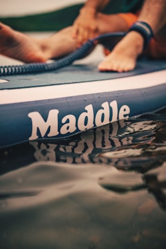 A close-up of a paddleboard with the brand name 'Maddle' visible. Feet are resting on the board, and a coiled leash is attached to an ankle. The water reflects the lettering on the board.