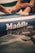 A close-up of a paddleboard with the brand name 'Maddle' visible. Feet are resting on the board, and a coiled leash is attached to an ankle. The water reflects the lettering on the board.