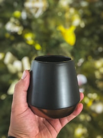 A hand is holding a sleek, black, modern cup against a blurred background that hints at a decorative or natural setting with greenery and bright colors.