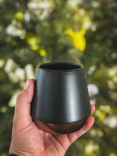 A hand is holding a sleek, black, modern cup against a blurred background that hints at a decorative or natural setting with greenery and bright colors.