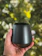 Close-up of a matte charcoal black Eco Flask held by a woman in a cozy café setting.