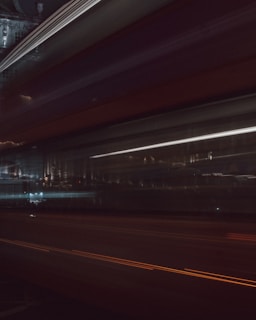 A blurred motion photograph capturing light trails and streaks against a dark urban backdrop. The image is characterized by dynamic horizontal lines, suggesting movement through a city at night. Faint reflections and architectural structures can be discerned in the background.