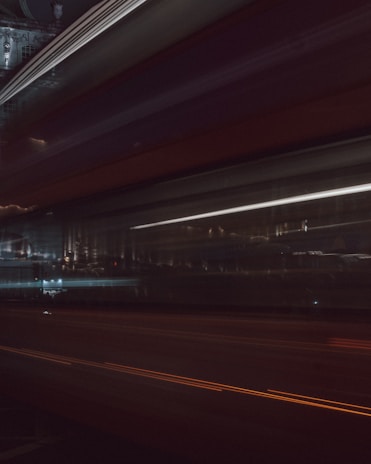 A blurred motion photograph capturing light trails and streaks against a dark urban backdrop. The image is characterized by dynamic horizontal lines, suggesting movement through a city at night. Faint reflections and architectural structures can be discerned in the background.