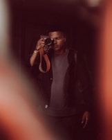 A person stands holding a camera, with one eye closed as they look through the viewfinder. The image is softly focused with warm, muted tones. The person wears a dark jacket over a gray shirt.