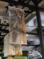 Hanging banners feature intricate botanical illustrations, including leaves and fruit. The artwork is monochromatic with orange accents, contrasting against the industrial ceiling with exposed beams and fixtures.