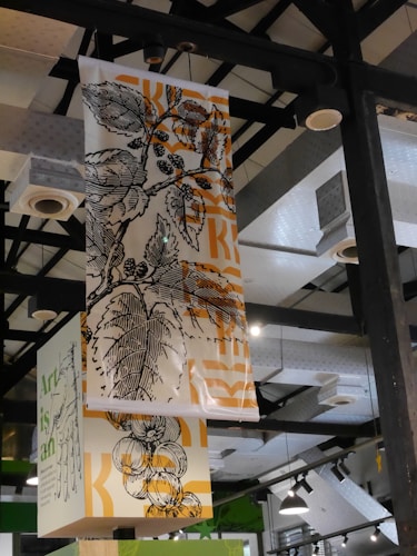 Hanging banners feature intricate botanical illustrations, including leaves and fruit. The artwork is monochromatic with orange accents, contrasting against the industrial ceiling with exposed beams and fixtures.