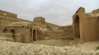 An ancient, partially ruined structure made of mud brick with arches and walls. The architecture suggests historical significance, surrounded by an arid landscape under an overcast sky.