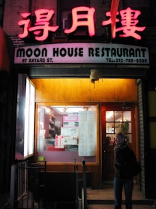 The entrance of a restaurant features a brightly lit neon sign with Chinese characters above an English sign that reads 'Moon House Restaurant'. The window displays various papers and posters, and there's a wooden door to the right. A person, wearing a scarf, stands near the entrance.