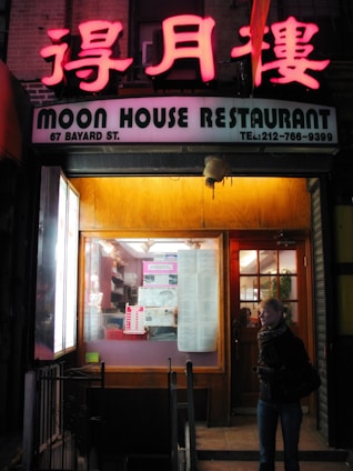 The entrance of a restaurant features a brightly lit neon sign with Chinese characters above an English sign that reads 'Moon House Restaurant'. The window displays various papers and posters, and there's a wooden door to the right. A person, wearing a scarf, stands near the entrance.