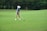 A young person wearing a blue striped shirt, beige shorts, white cap, and golf gloves is preparing to hit a golf ball on a lush green golf course. The scene is surrounded by trees in the background, providing a serene and natural setting.