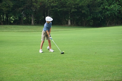 A young person wearing a blue striped shirt, beige shorts, white cap, and golf gloves is preparing to hit a golf ball on a lush green golf course. The scene is surrounded by trees in the background, providing a serene and natural setting.