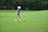 A young person wearing a blue striped shirt, beige shorts, white cap, and golf gloves is preparing to hit a golf ball on a lush green golf course. The scene is surrounded by trees in the background, providing a serene and natural setting.