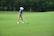 A young person wearing a blue striped shirt, beige shorts, white cap, and golf gloves is preparing to hit a golf ball on a lush green golf course. The scene is surrounded by trees in the background, providing a serene and natural setting.