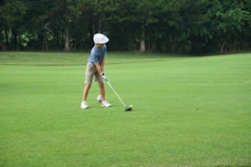 A young person wearing a blue striped shirt, beige shorts, white cap, and golf gloves is preparing to hit a golf ball on a lush green golf course. The scene is surrounded by trees in the background, providing a serene and natural setting.